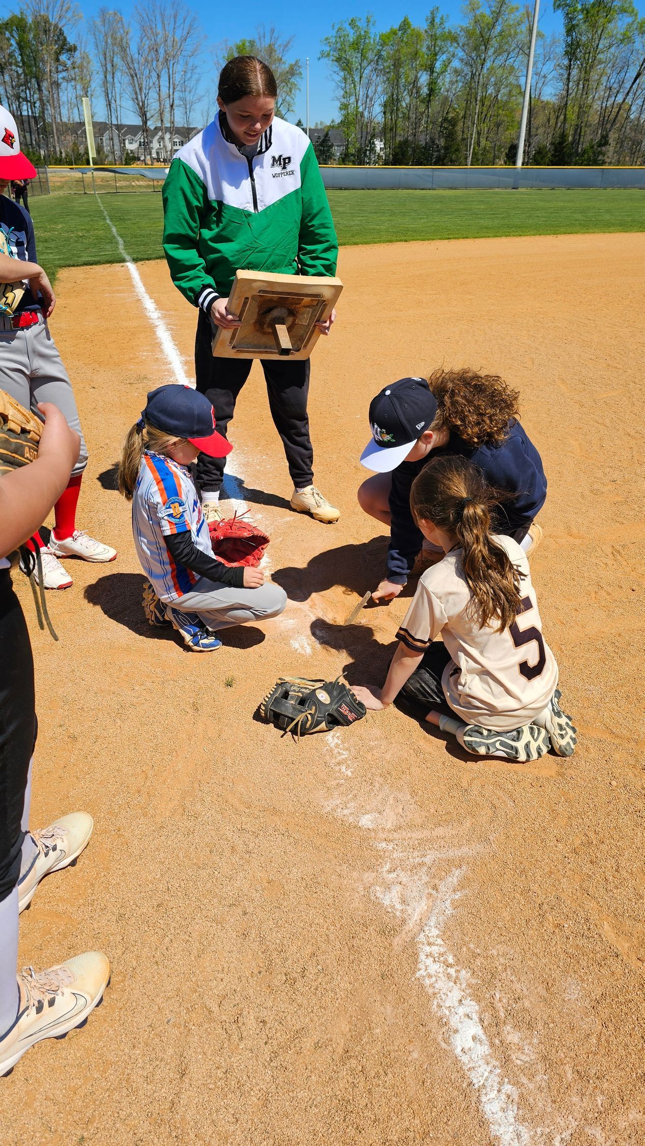 Children crouch on a baseball field while an adult holds a base tray, with a white line marked on the dirt.