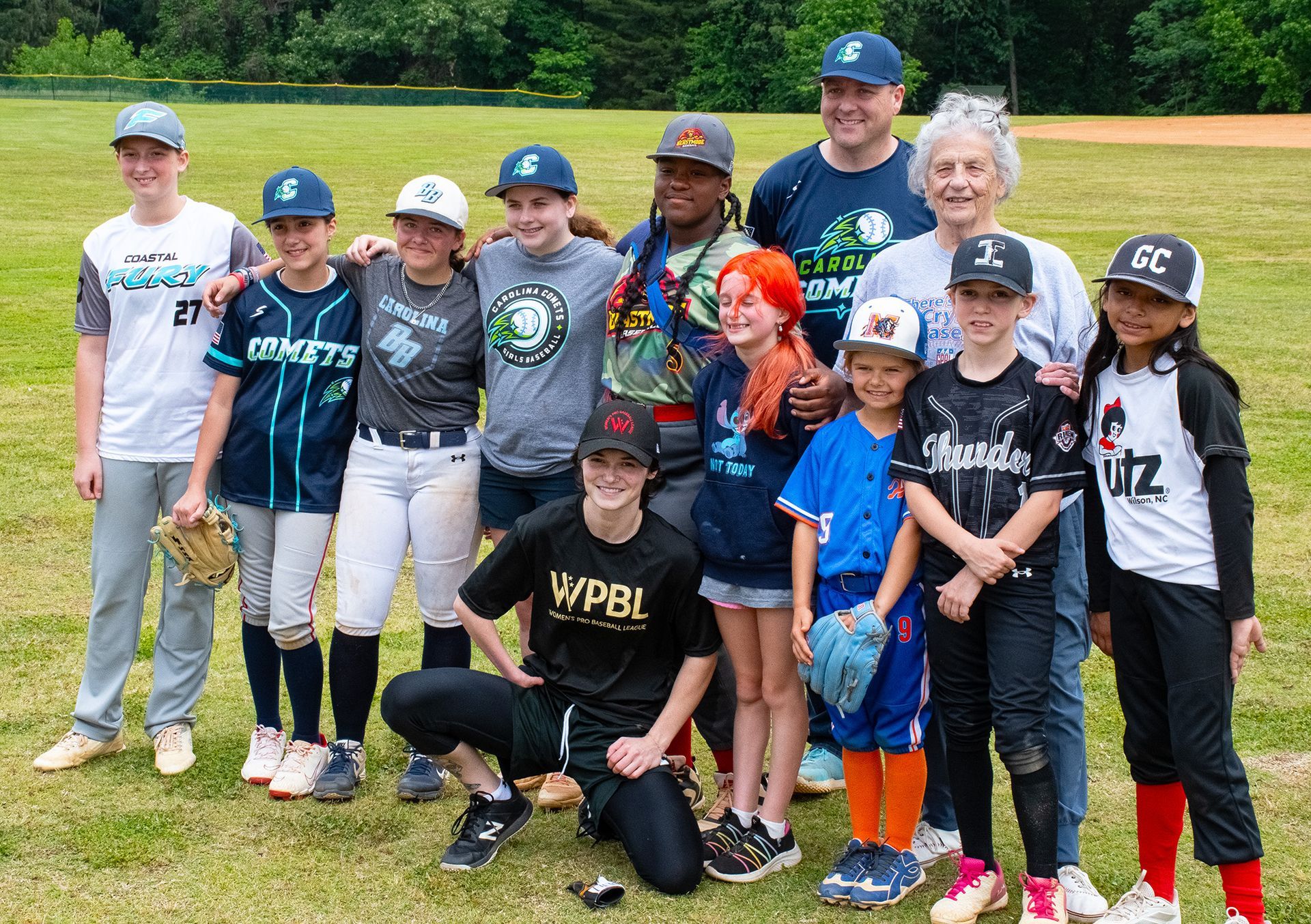 Girls baseball team posing on a field with coaches, smiling in uniforms and caps