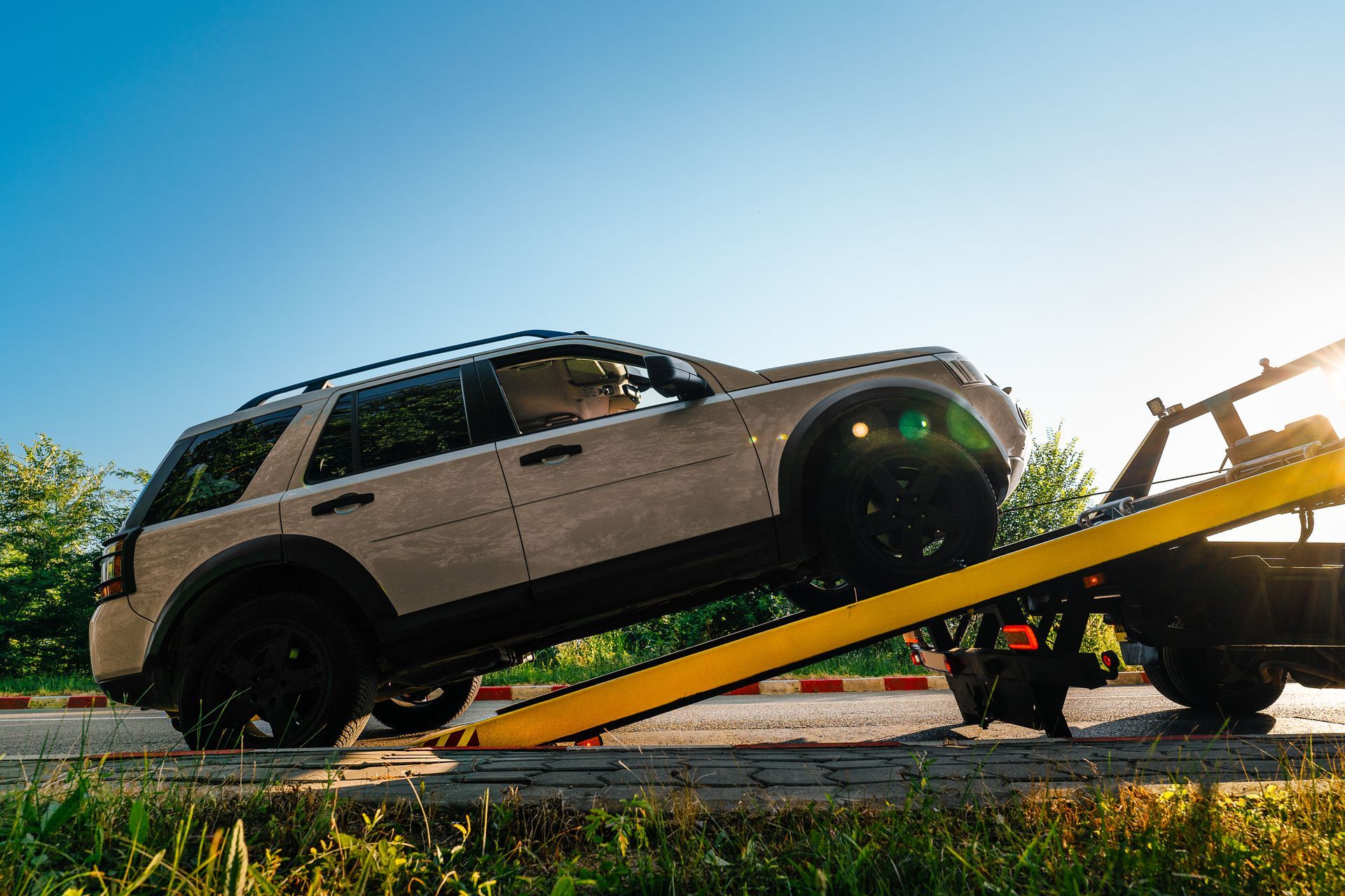 A light-colored SUV is being winched up onto the yellow flatbed of a tow truck on a sunny day. | John’s Auto Center
