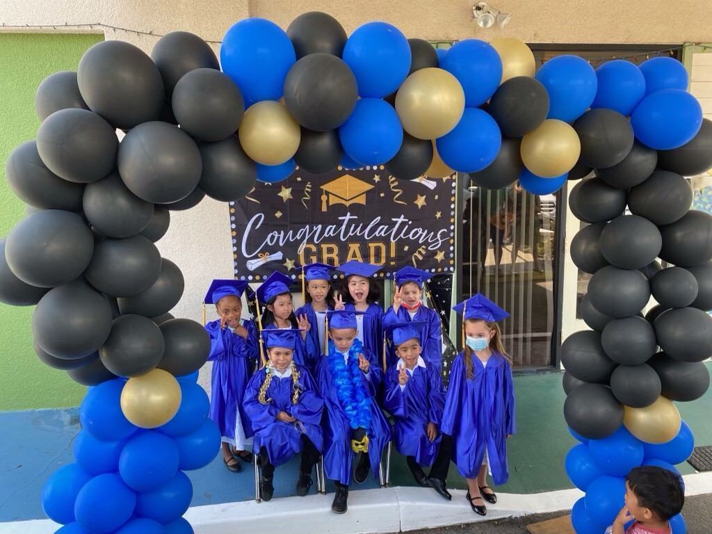 A group of children in graduation caps and gowns are standing under a balloon arch.