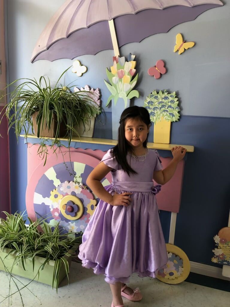 A little girl in a purple dress is standing under an umbrella.