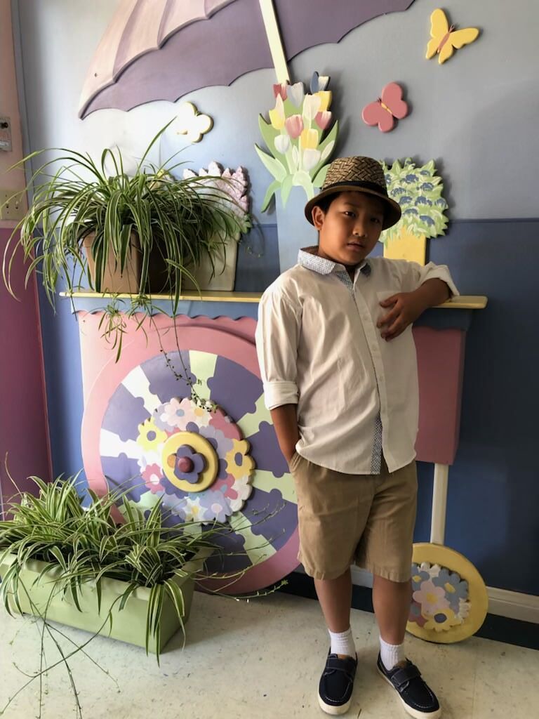 A young boy wearing a hat is standing in front of a cart with plants on it.