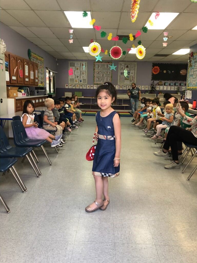 A little girl in a blue dress is standing in a classroom.