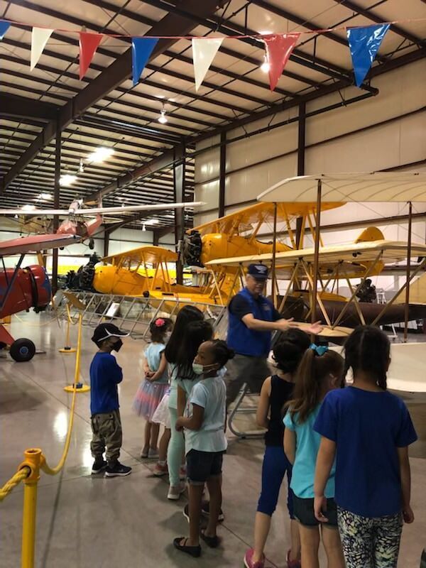 A group of children are standing in front of planes in a hangar.
