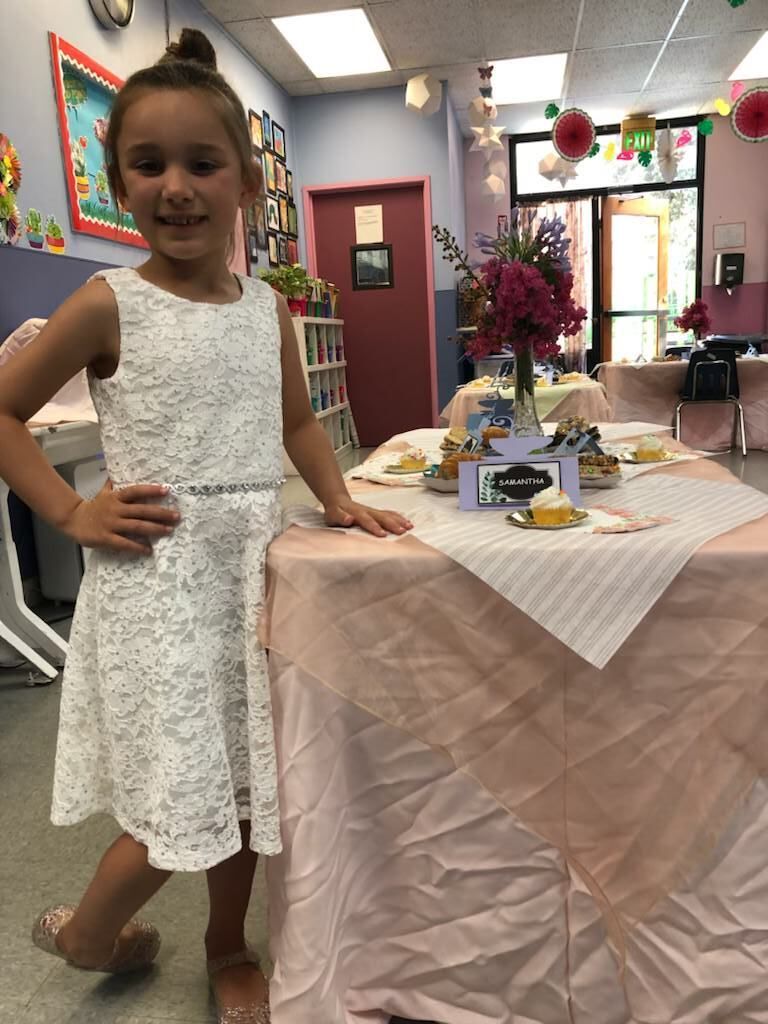 A little girl in a white dress is standing next to a table
