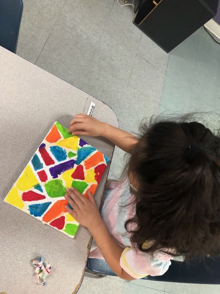A little girl is sitting at a table making a colorful painting.