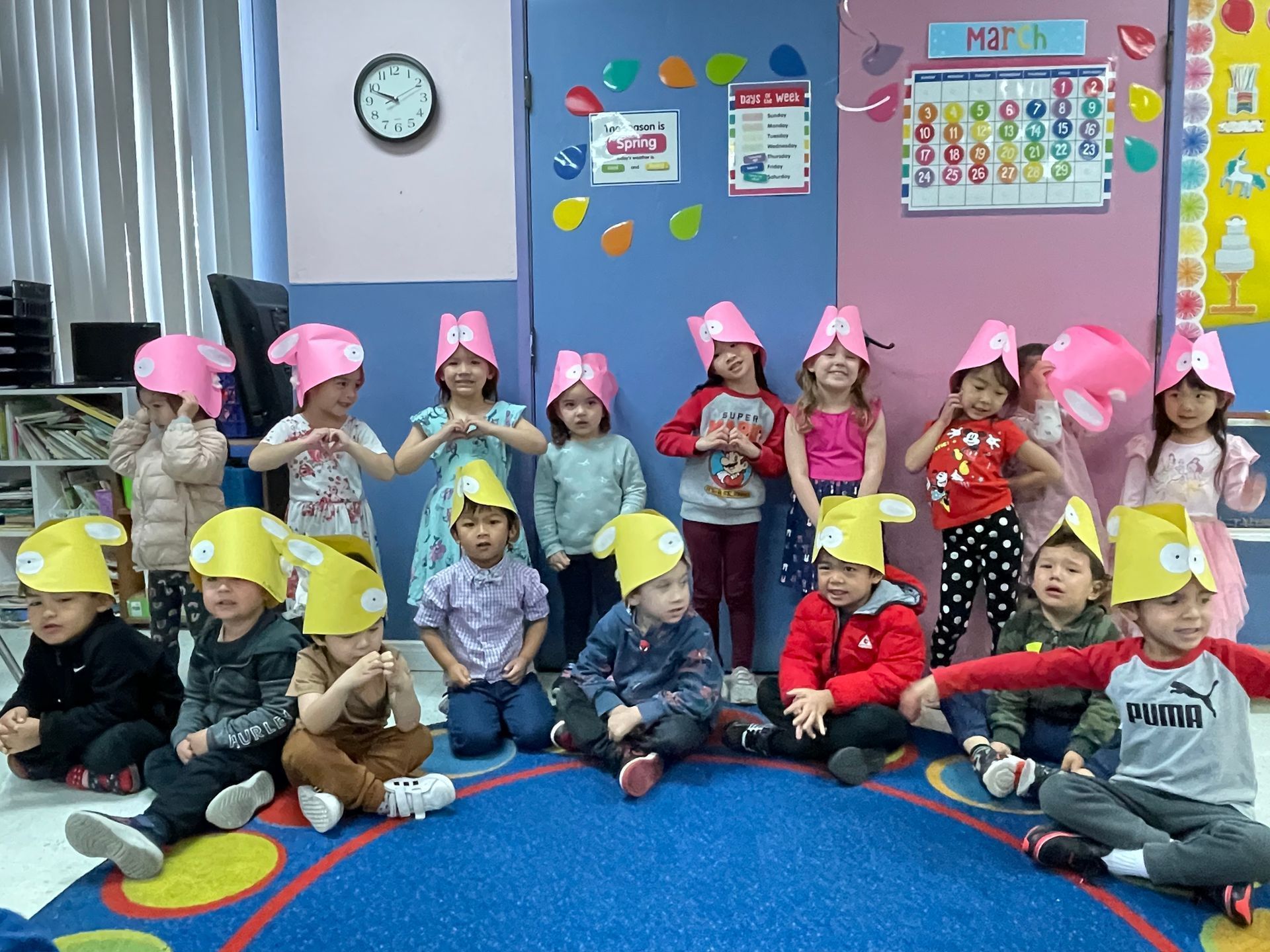 A group of children wearing paper hats are sitting on the floor in a classroom.