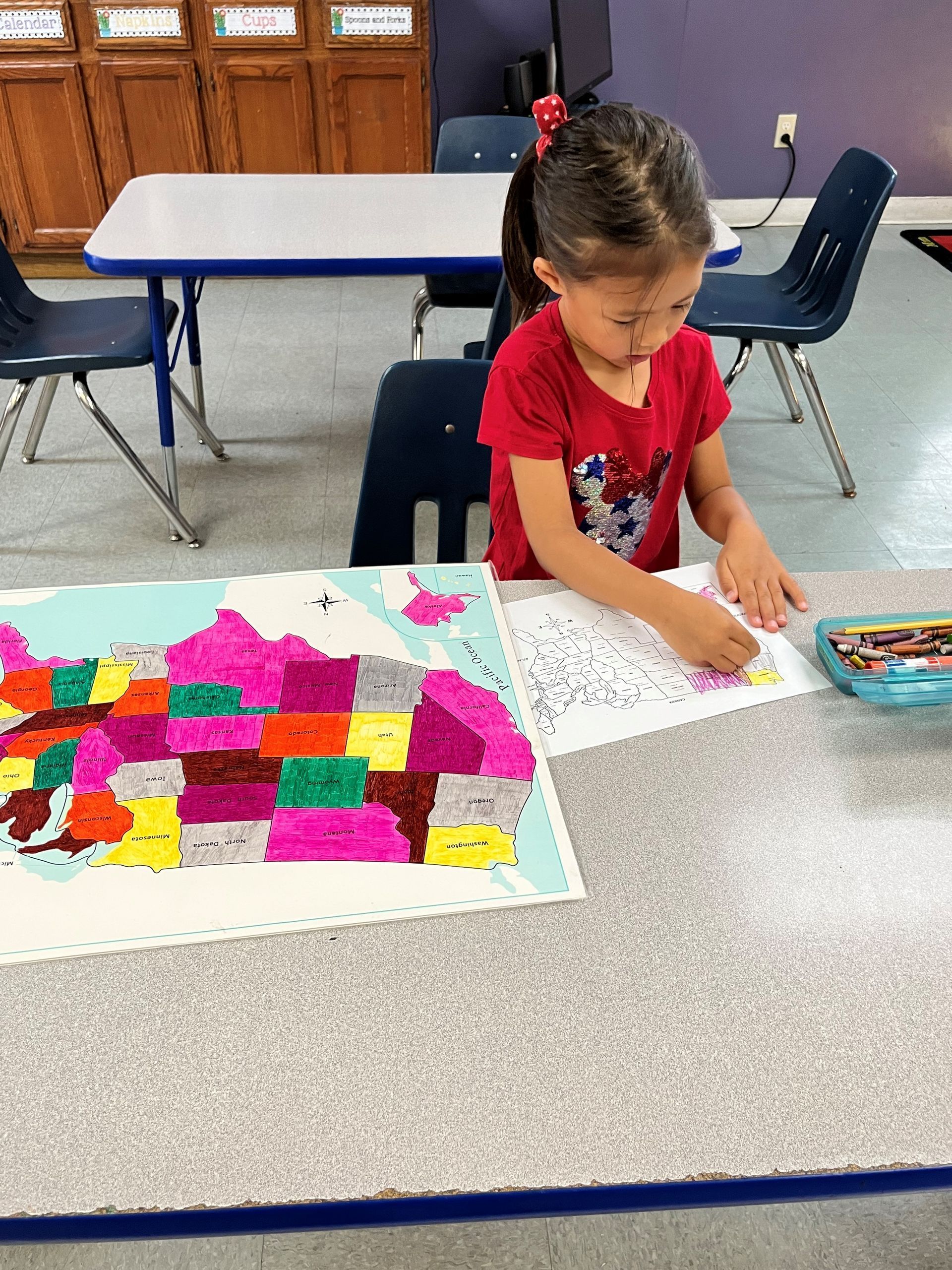 A little girl is sitting at a table painting a map of the united states.