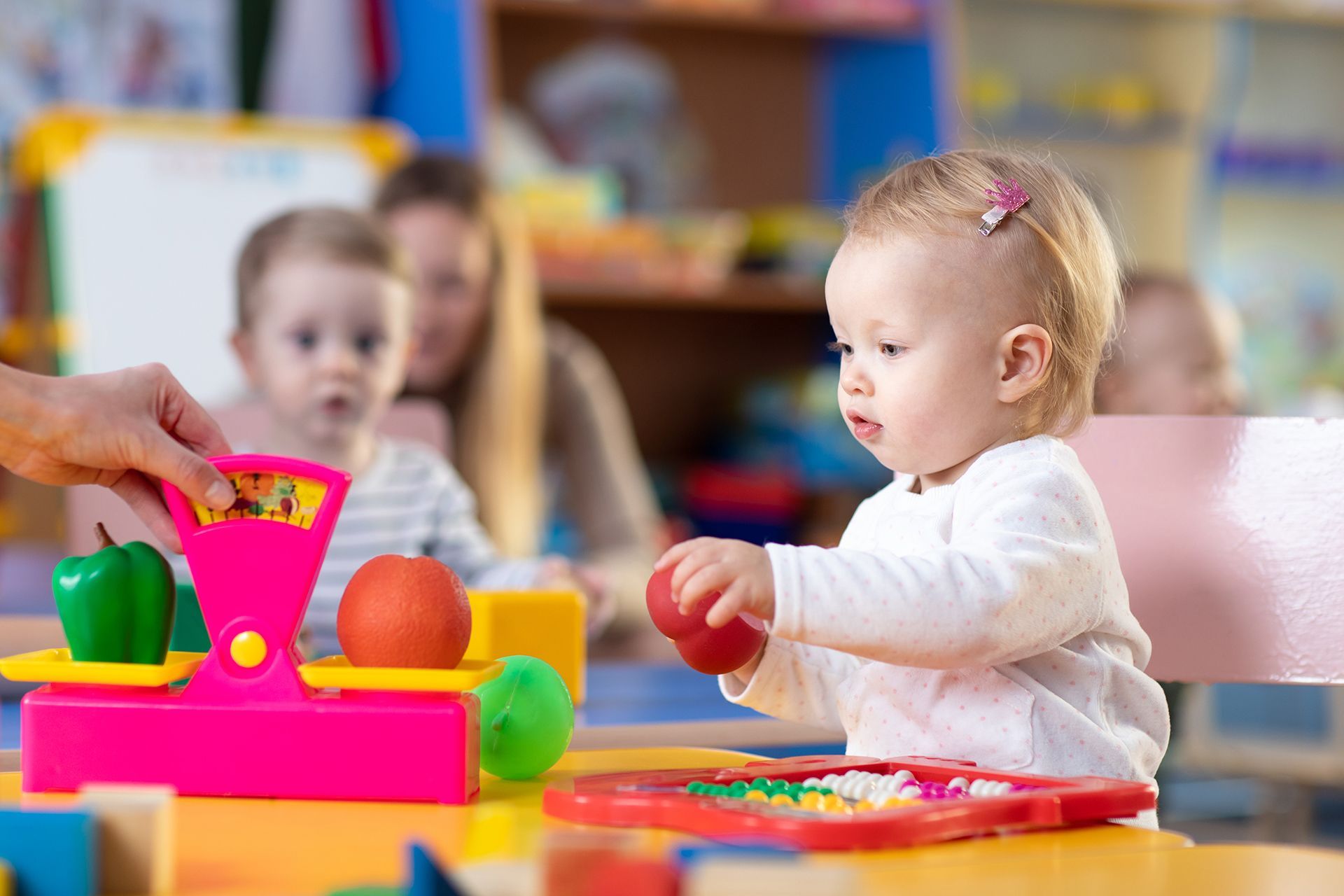 A baby is playing with a toy scale in a classroom.