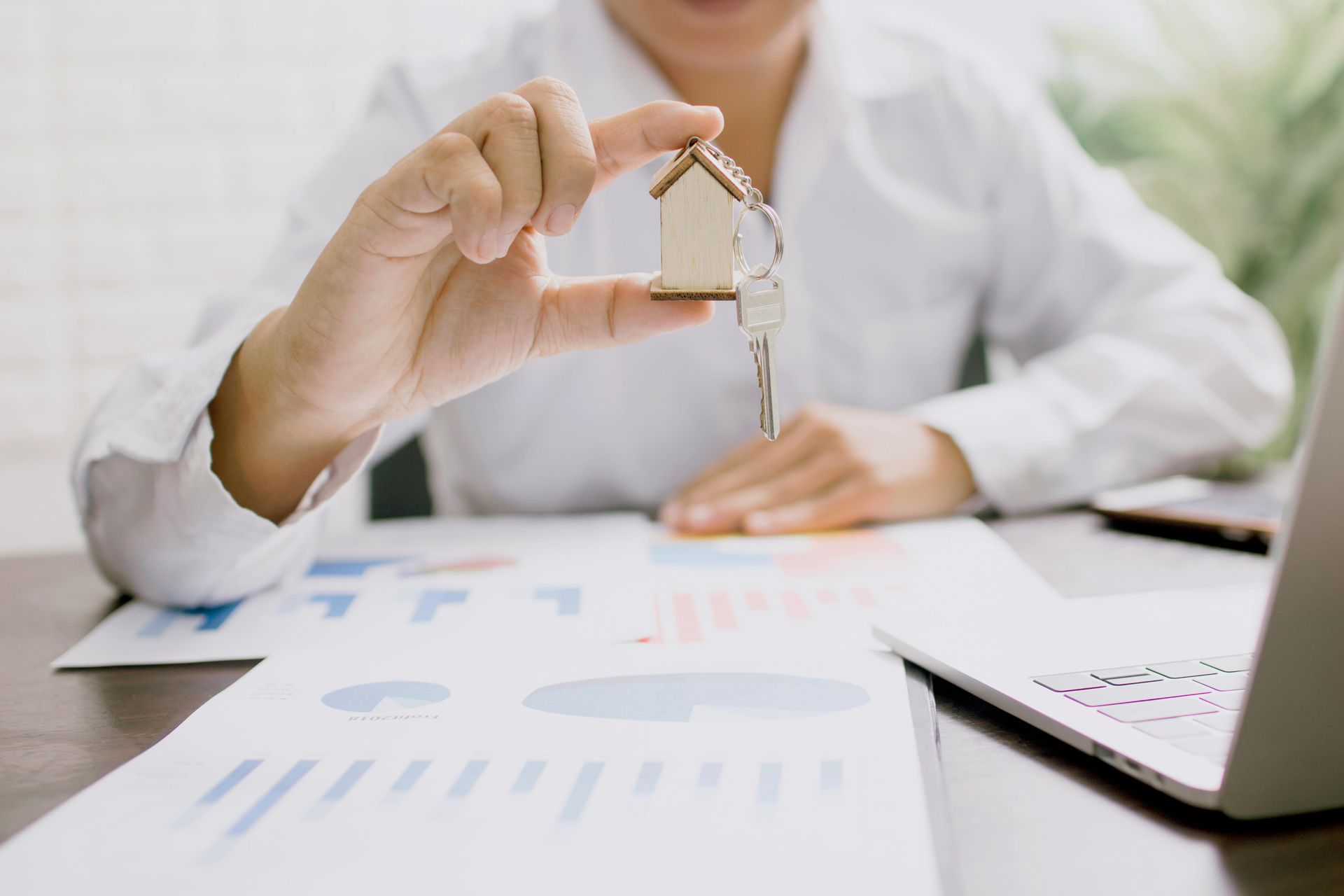 A woman is sitting at a desk holding a small house and keys.