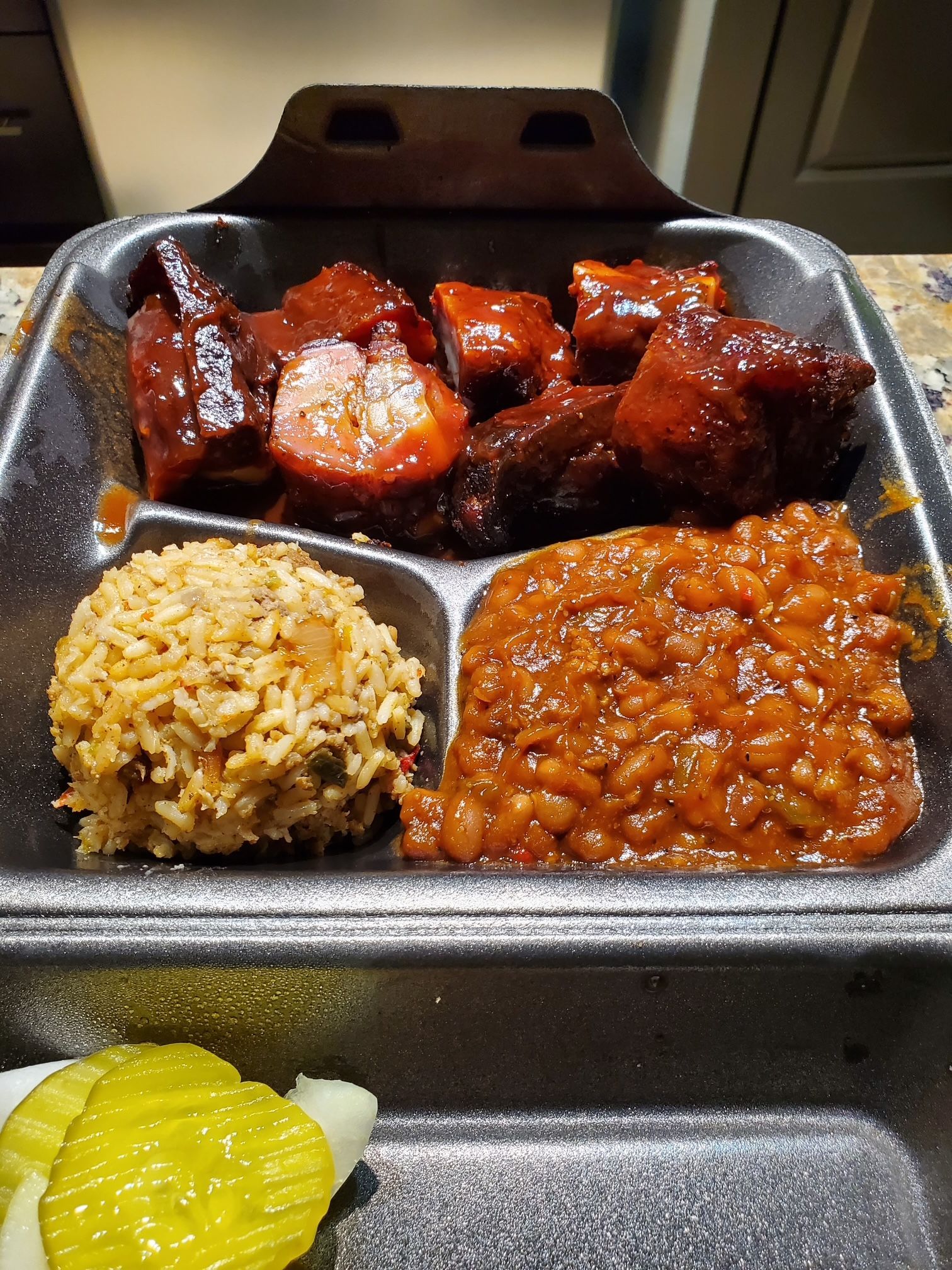 A styrofoam tray filled with meat , beans , rice and pickles.