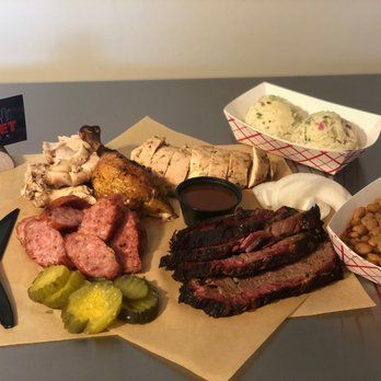 BBQ platter with sliced brisket, sausage, chicken, pickles, beans, potato salad, and sauce.