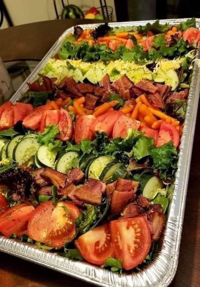 A large tray of salad with tomatoes , cucumbers , carrots and bacon.