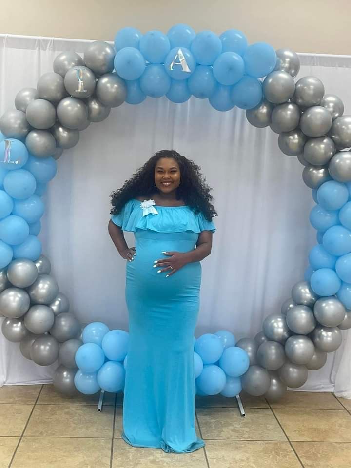 A pregnant woman in a blue dress is standing in front of a balloon arch.