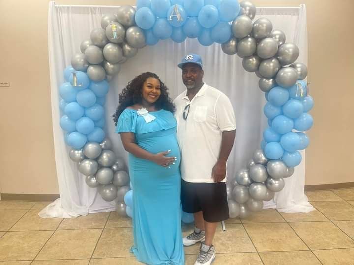 A man and a pregnant woman are standing in front of a balloon arch.