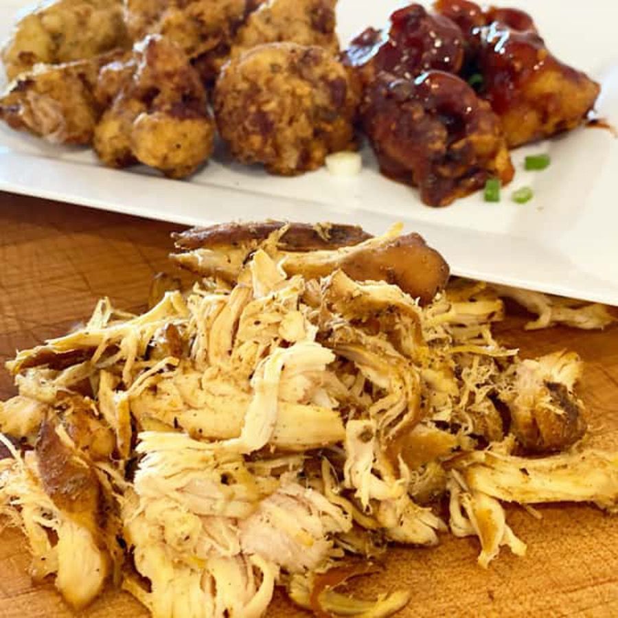Shredded chicken is sitting on a wooden cutting board next to a plate of chicken wings.
