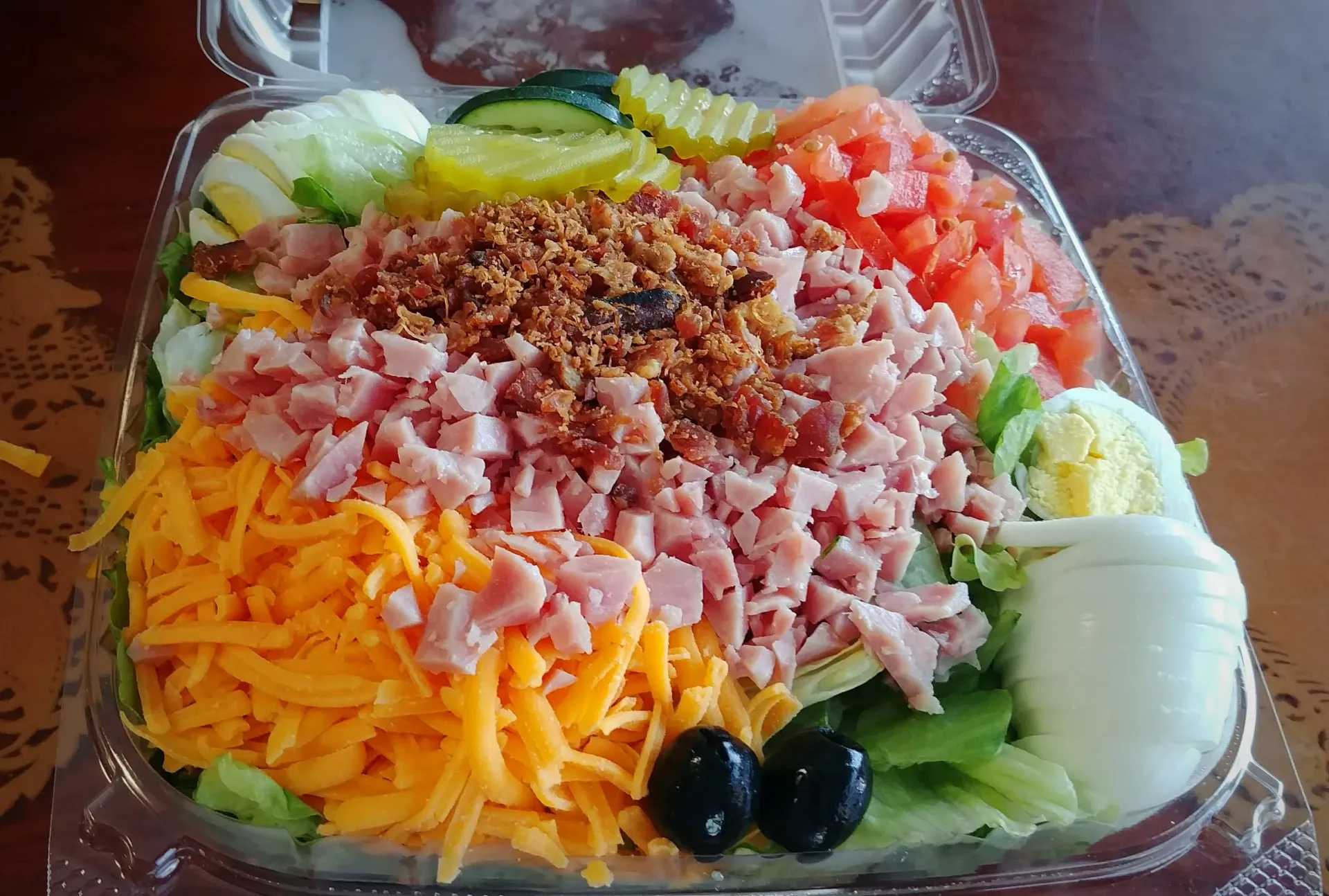 A plastic container filled with a salad with ham , cheese , eggs , pickles and olives.
