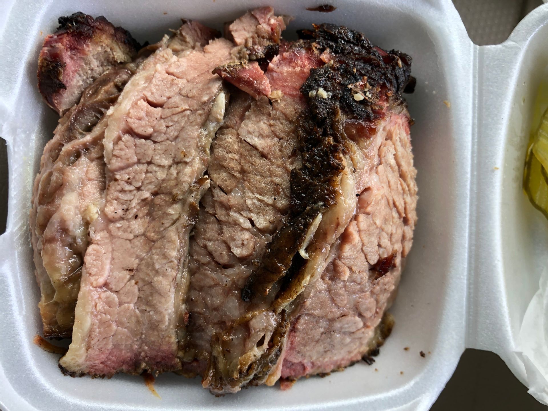 A styrofoam container filled with sliced meat next to a bowl of pickles.
