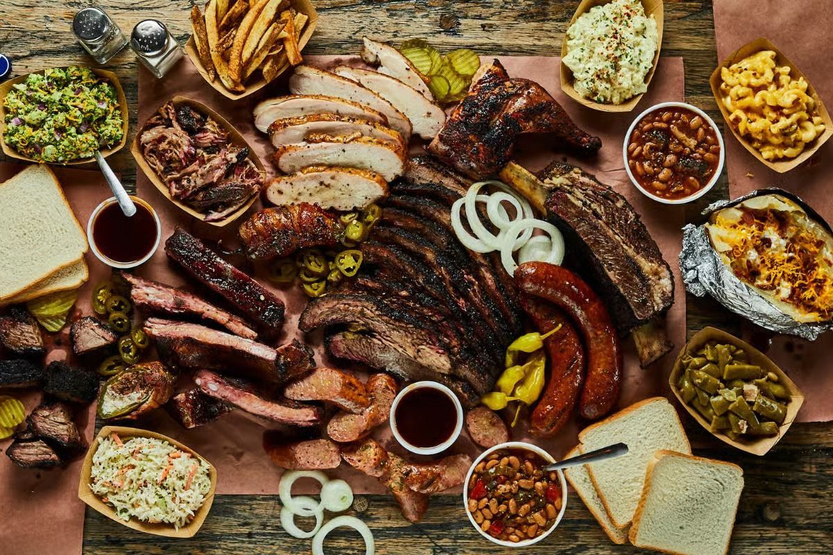 Overhead view of a BBQ spread with various meats, sides, sauces, and bread on a wooden surface.