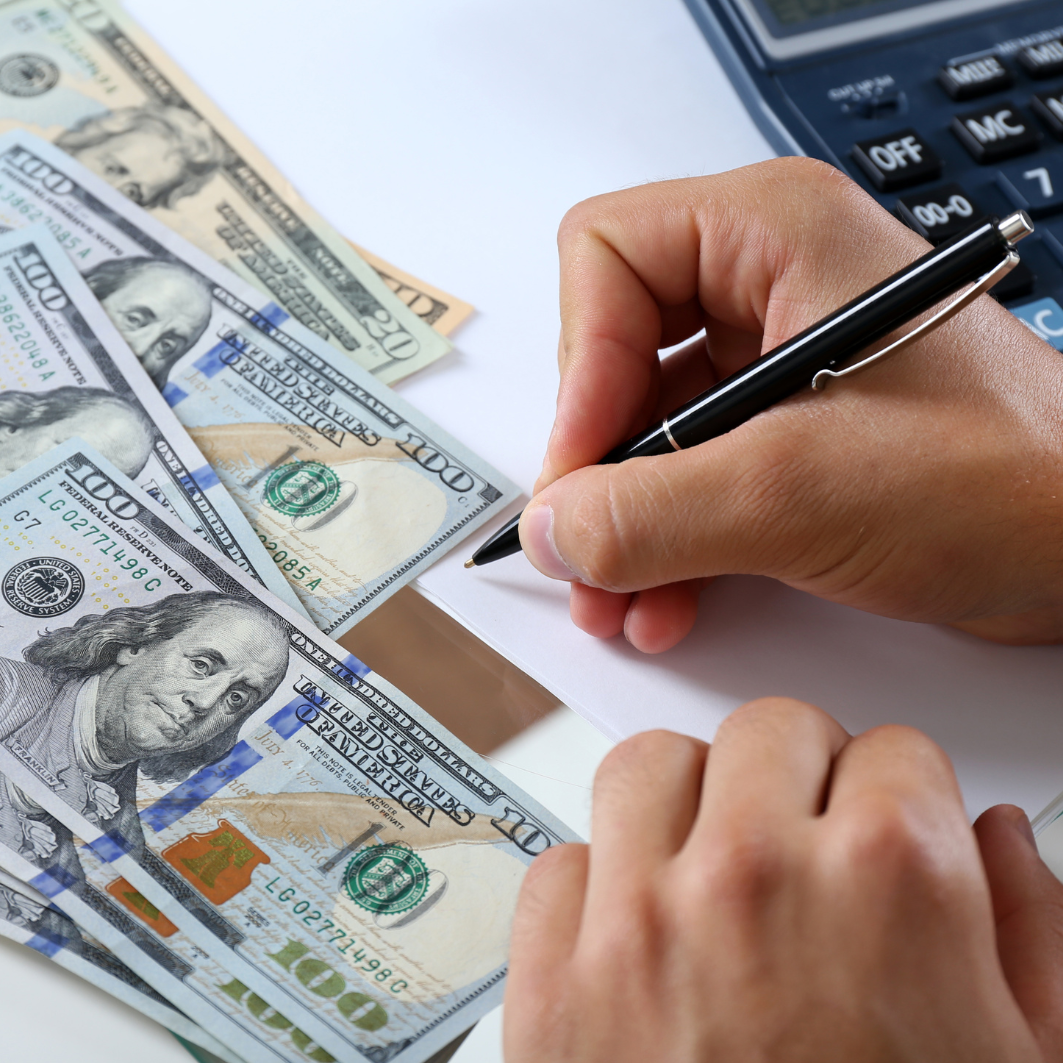 A person is writing on a piece of paper in front of a pile of 100 dollar bills