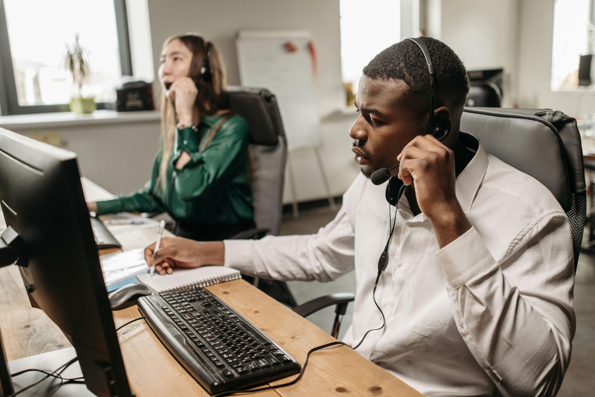A man and a woman are sitting at a desk in front of a computer talking on a headset.