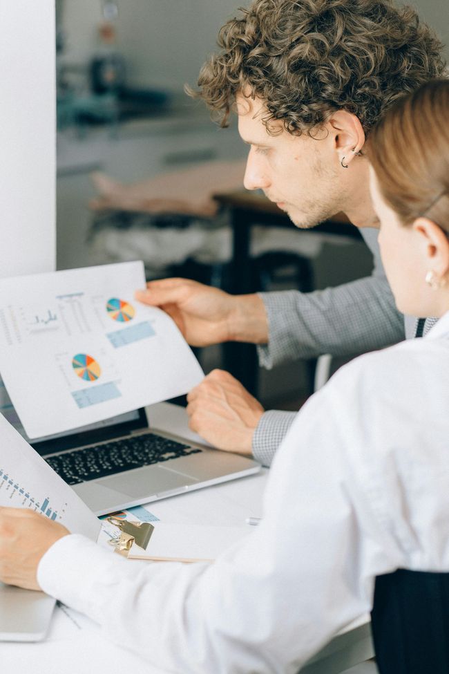 A man and a woman are sitting at a desk looking at a laptop.