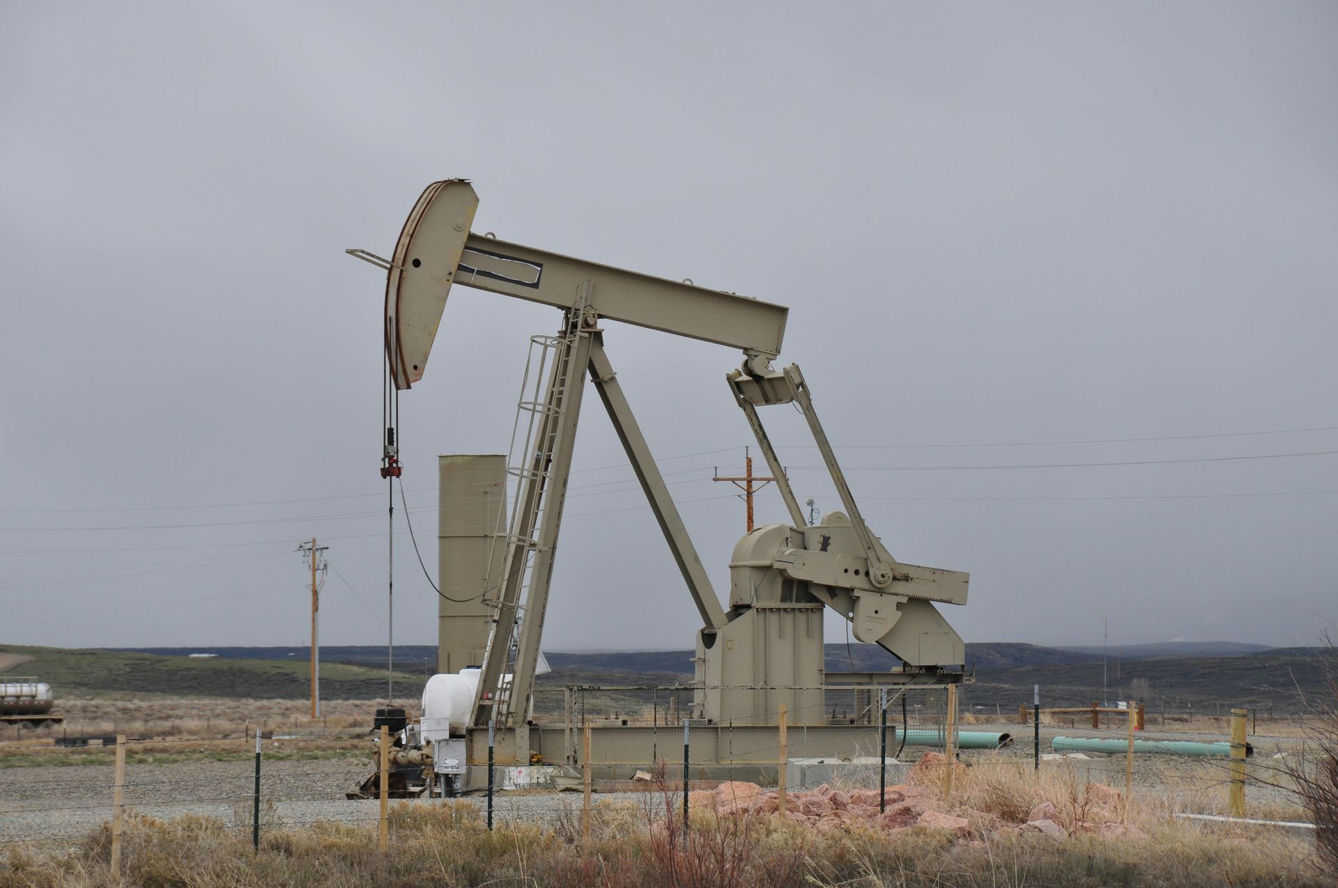 Oil pumpjack in a barren field under a gray sky, with pipes and fencing in the foreground.