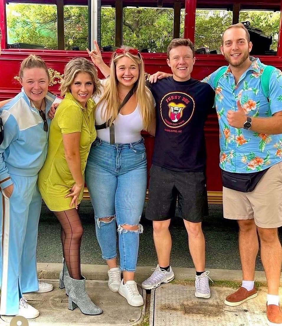 Five friends pose together in front of a red trolley, smiling and giving a thumbs up.