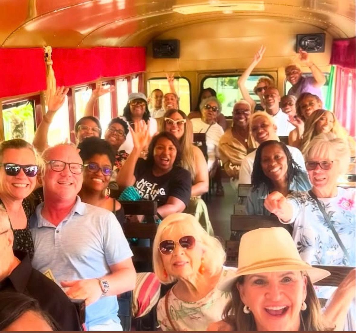 A large group of people smiling and waving while riding on a bus with red interior accents.