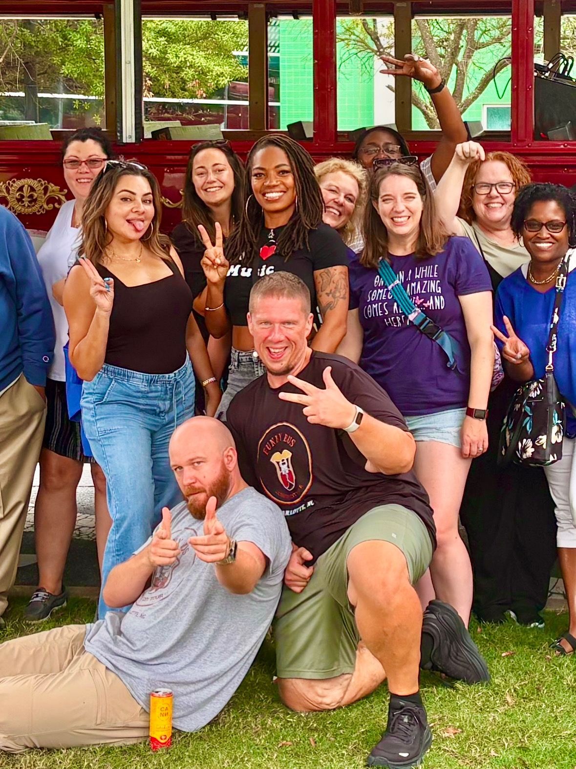 A diverse group poses in front of a red trolley; two people in the front kneel and make hand gestures toward the camera.