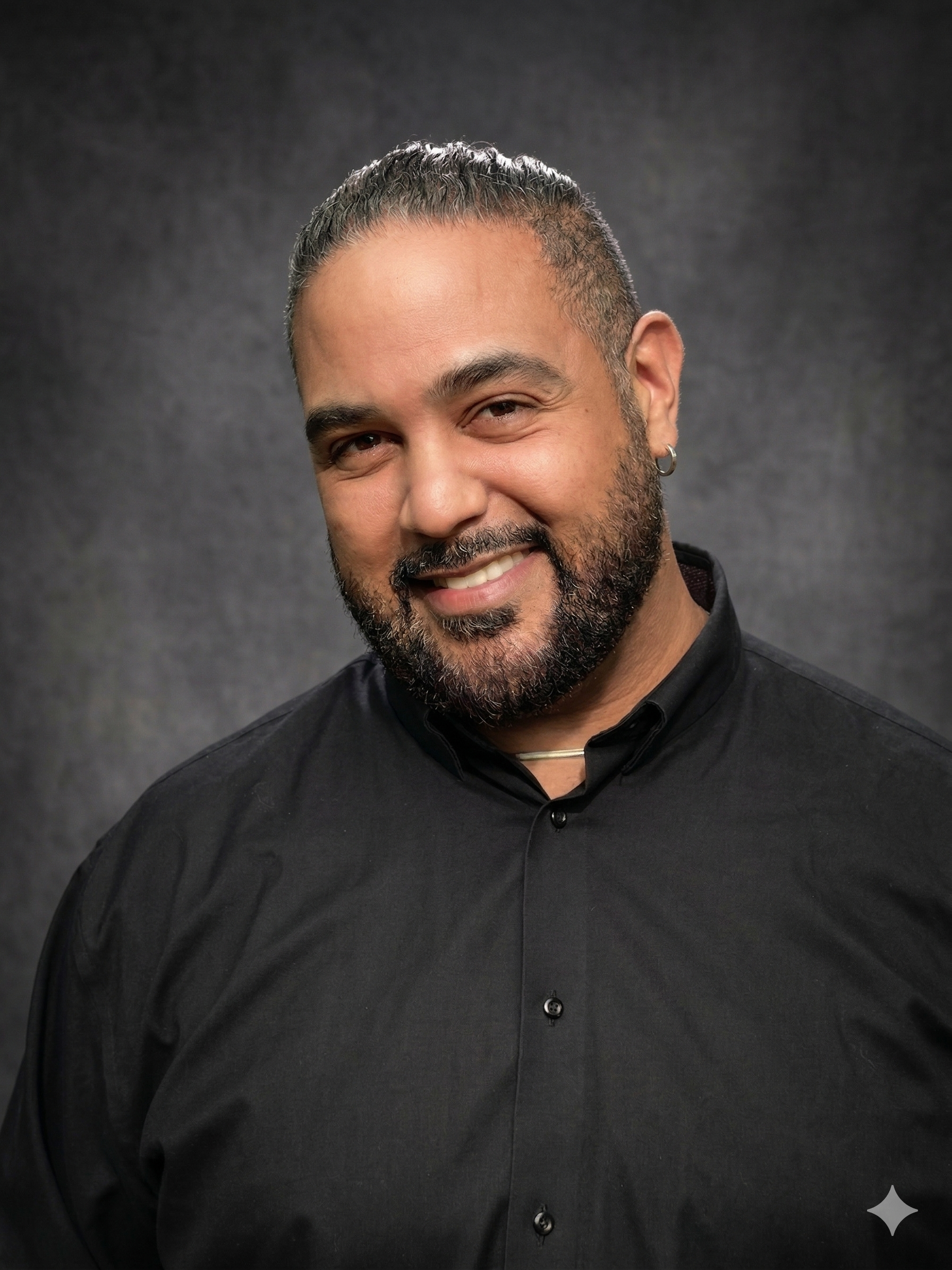 Man in white shirt touches his head, smiling at the camera, against a dark background.