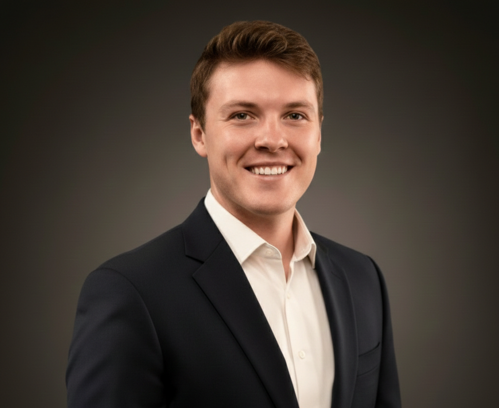 Man in a navy blazer and white shirt smiles at the camera against a gray background.