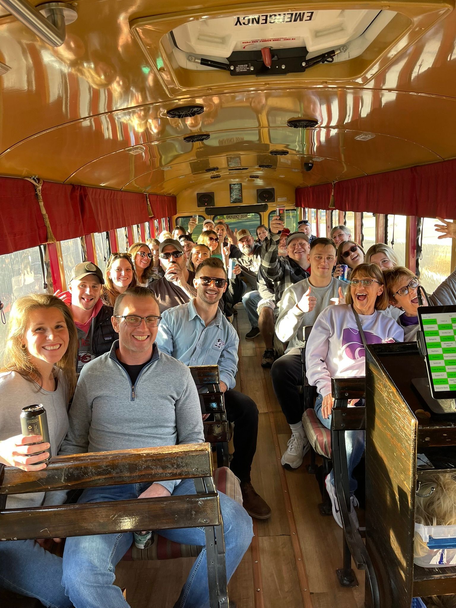 People on a vintage bus, smiling. Interior shot with wooden benches, red curtains, and decorative lighting.