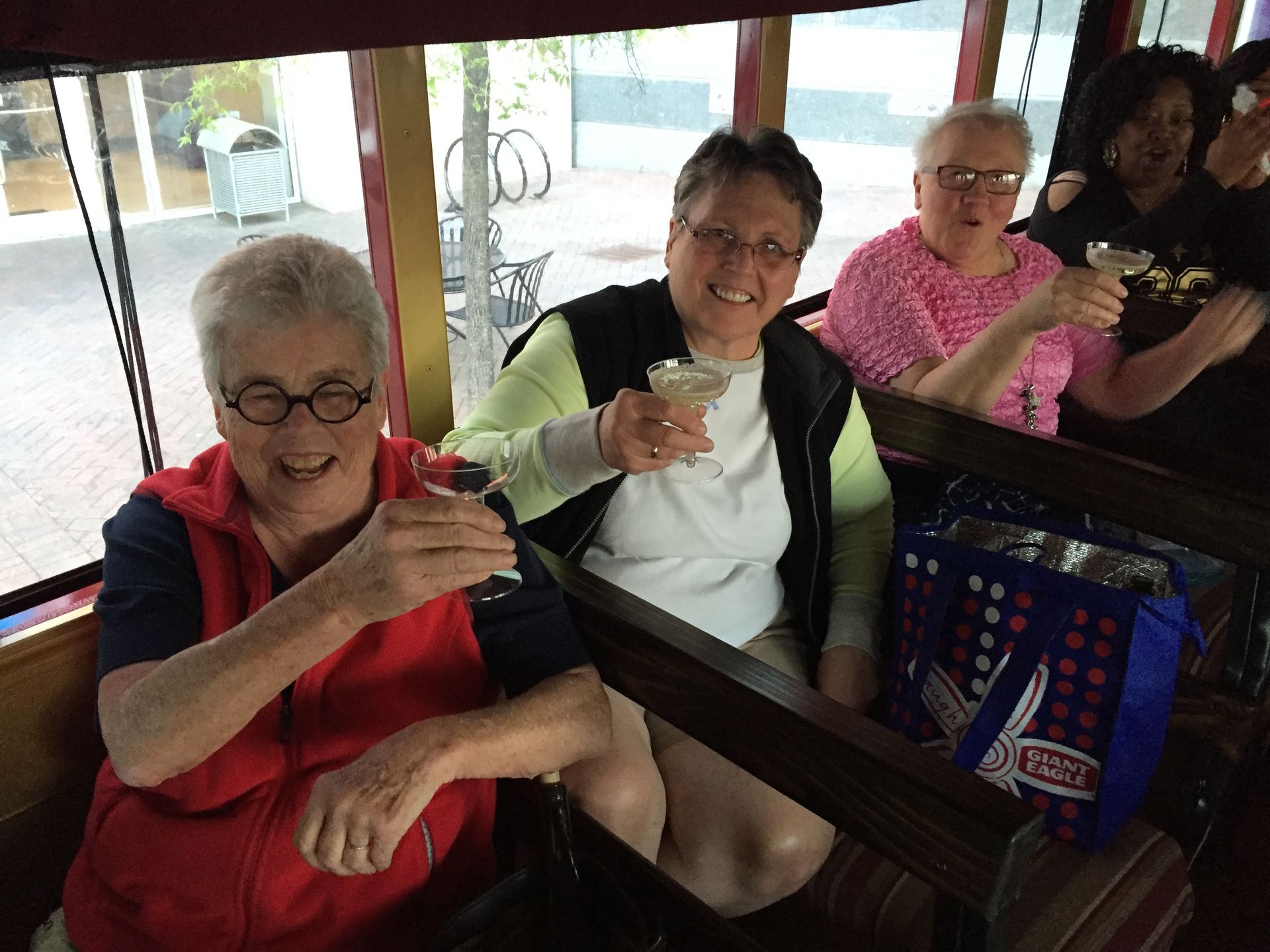 Three smiling people sitting in a trolley raise cocktail glasses in a toast, with a blue patterned tote bag on a seat.