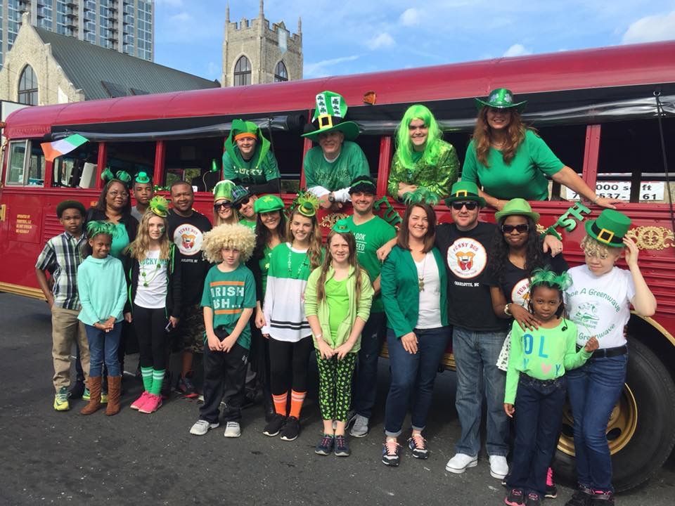A group posing for a photo in front of a red bus, wearing green attire and St. Patrick's Day themed accessories.