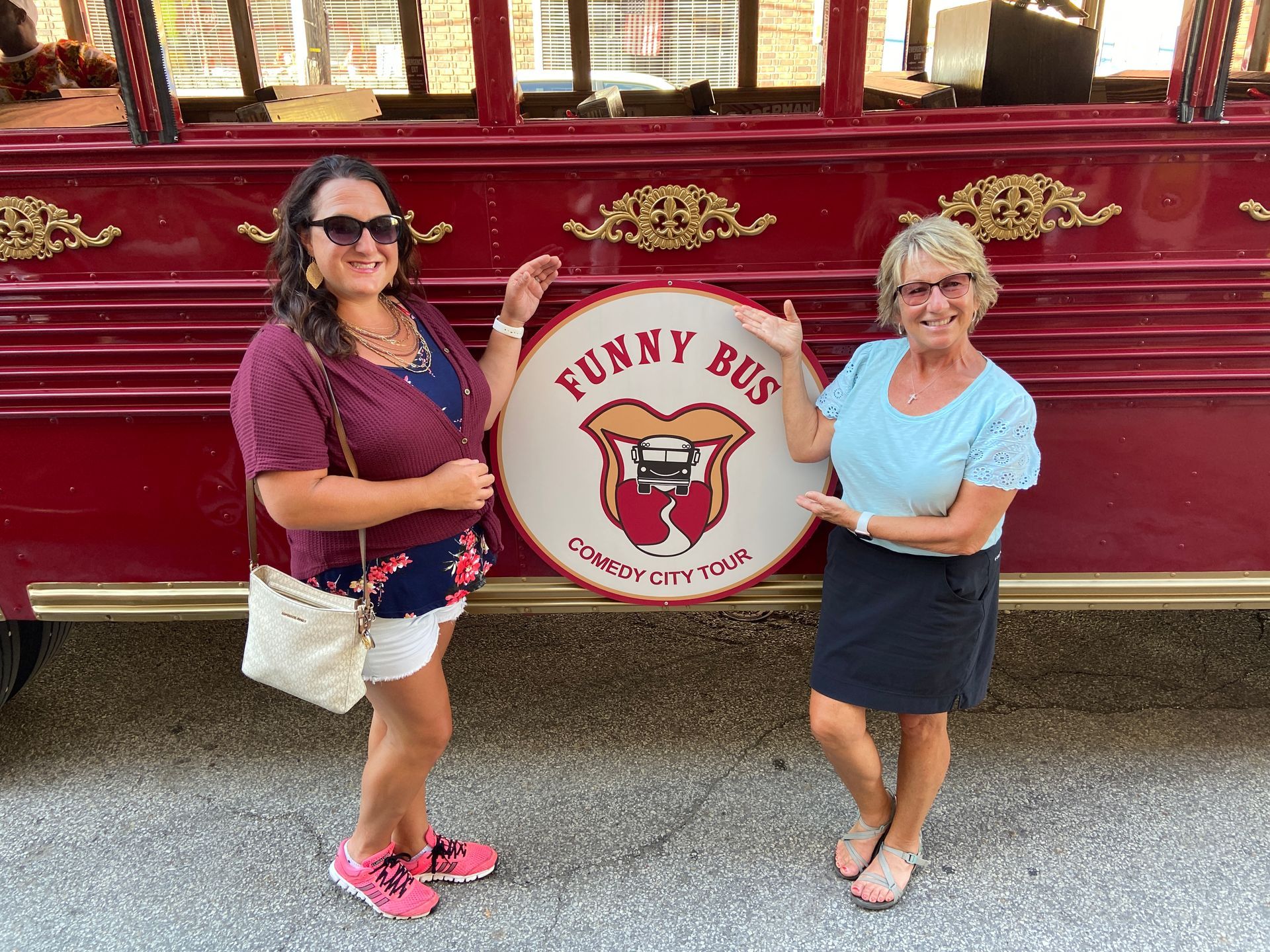Two smiling people stand in front of a red Funny Bus tour vehicle, gesturing toward the circular company logo.