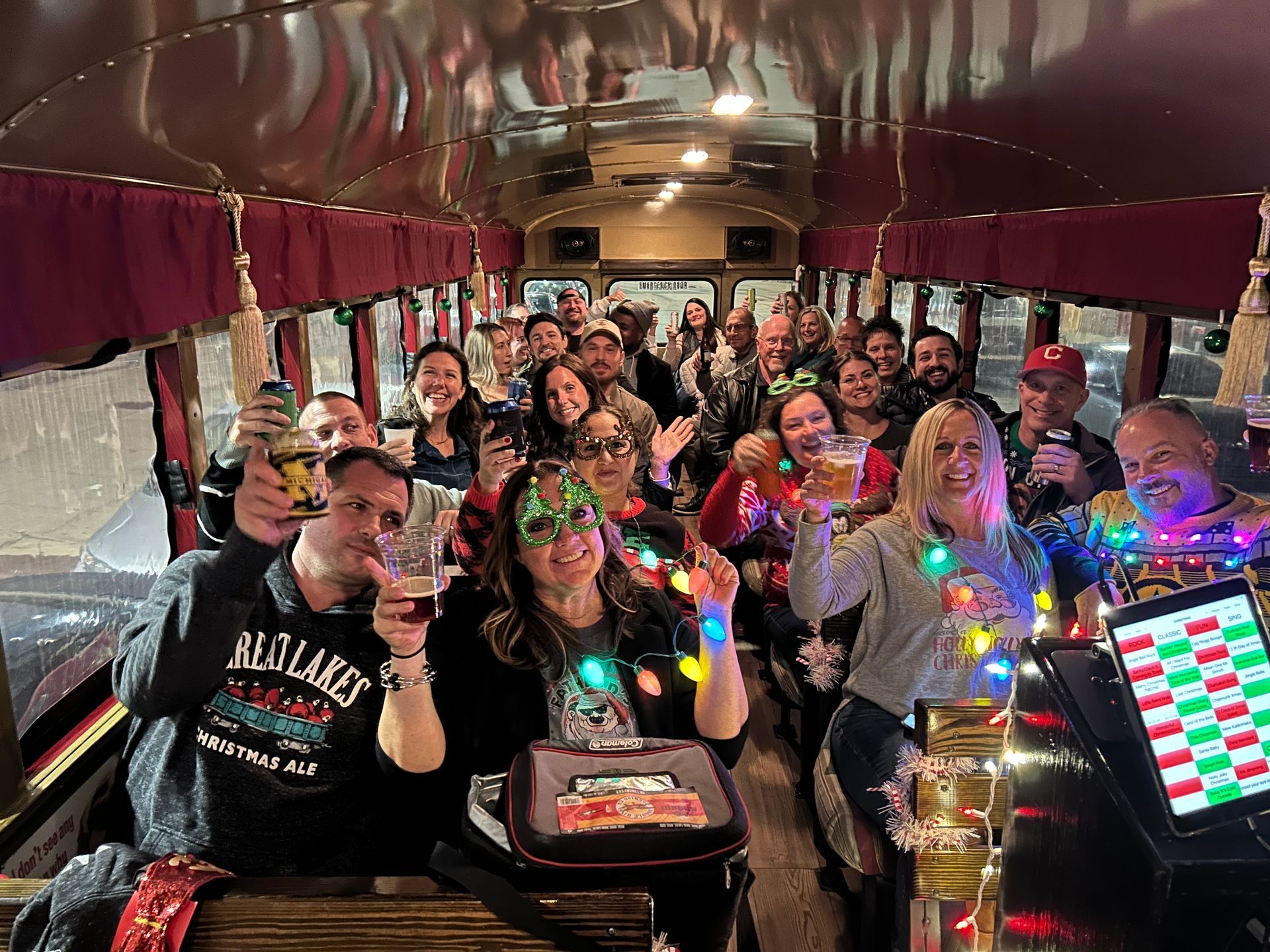 Group of people smiling inside a party bus, wearing colorful lights and holding drinks and a cake