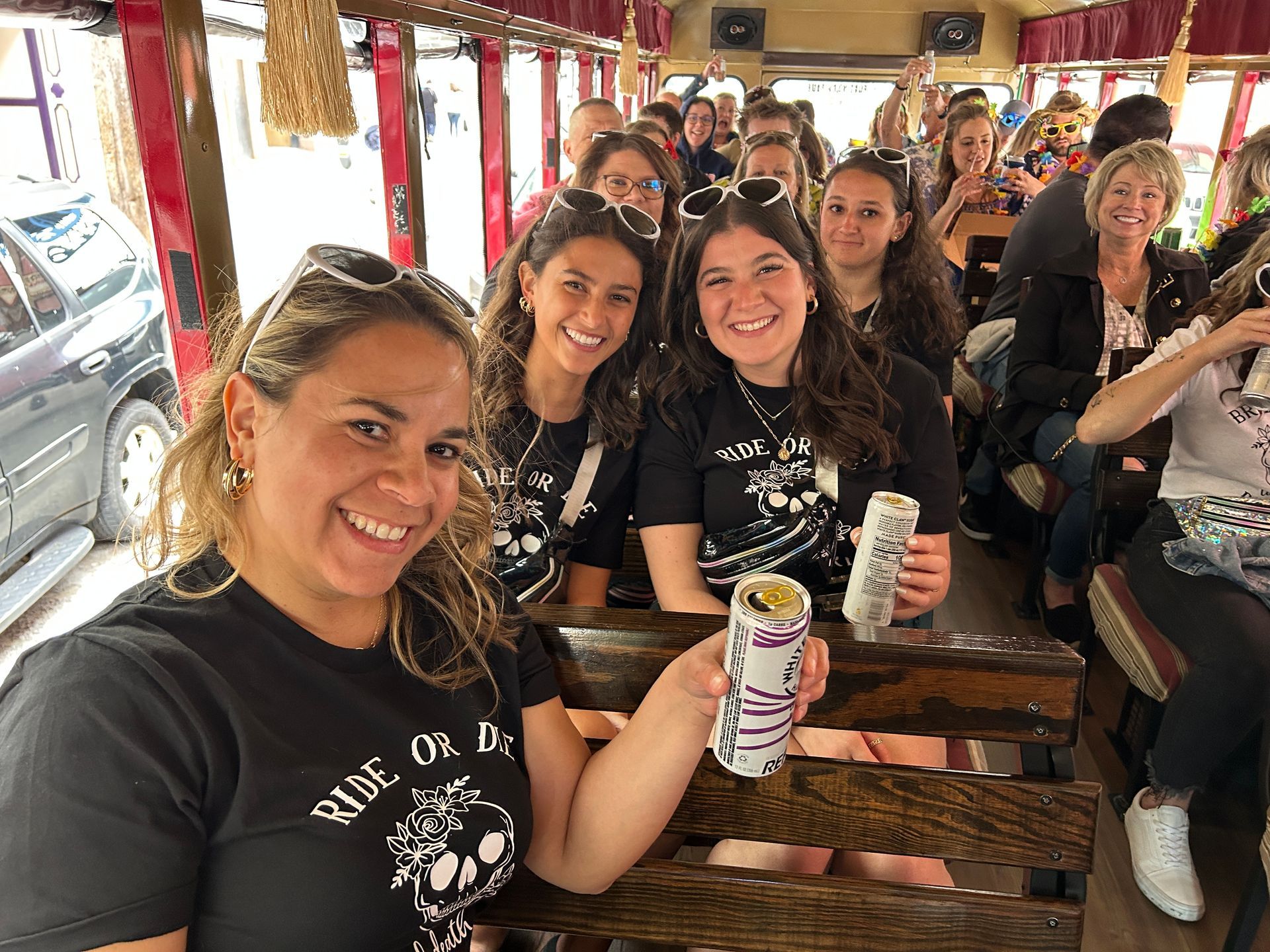 A group of people wearing matching black shirts smile while riding on an open-air trolley, some holding canned drinks.