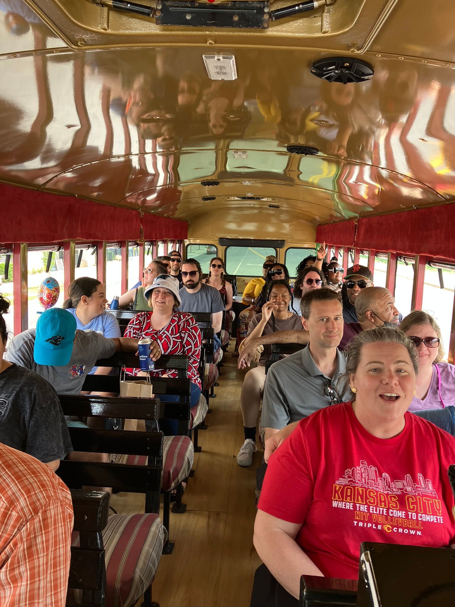 A group of people sits inside a bus with reflective, gold-toned ceilings, many smiling at the camera.