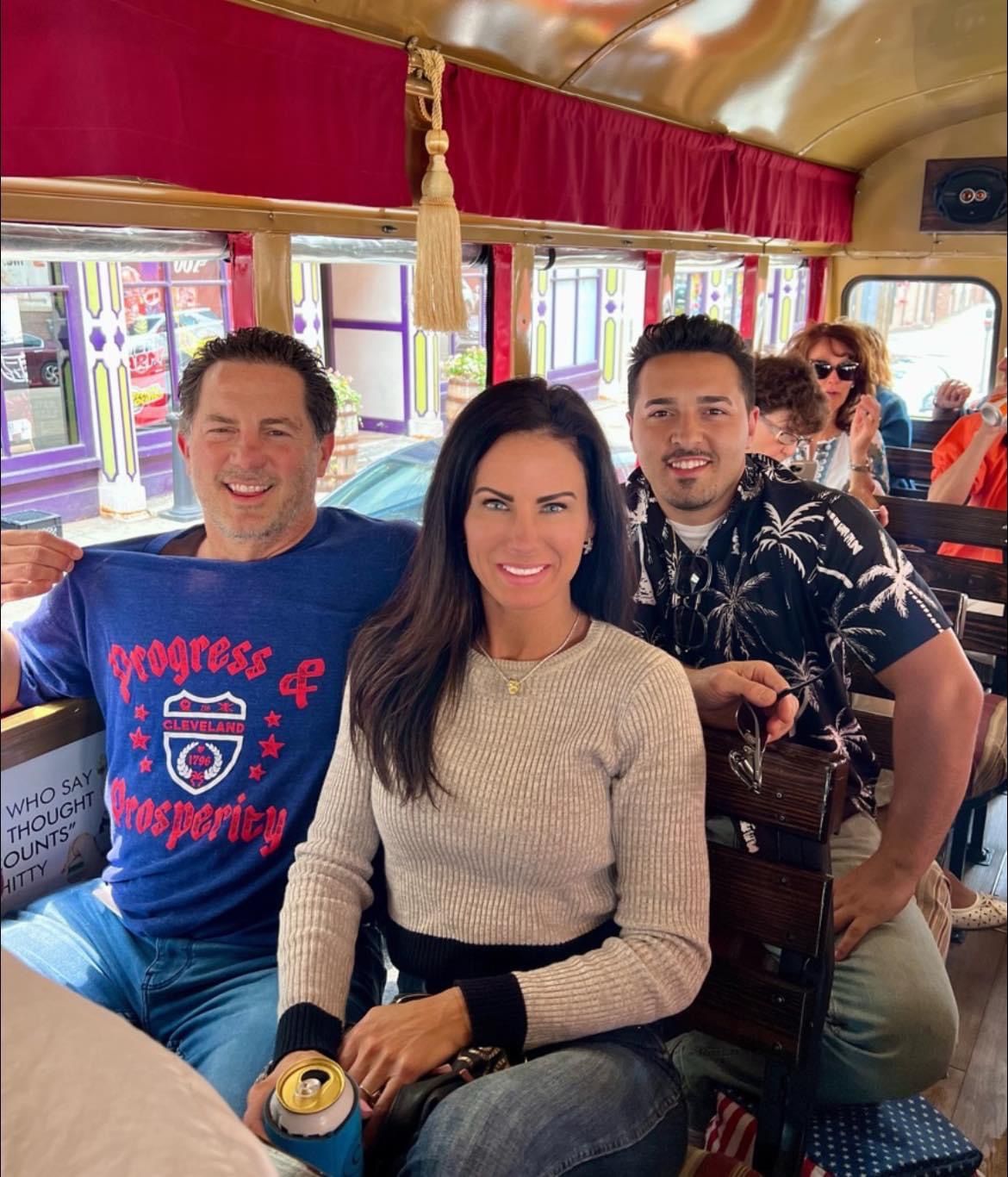 Three people sit in the interior of a trolley car; one person holds a beverage and another wears a blue graphic shirt.