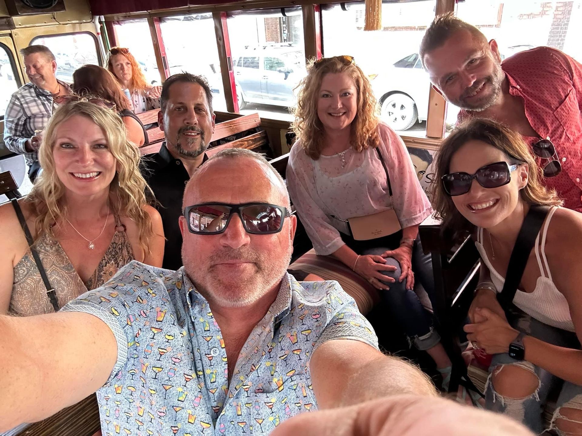 Group of six smiling people taking a selfie inside a vehicle, with bright daylight outside.