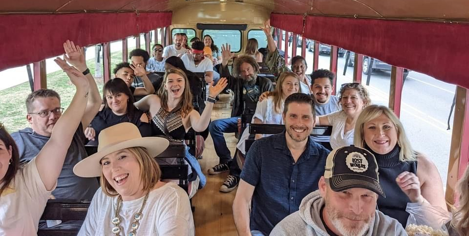Passengers smiling and waving inside a trolley bus with red curtains