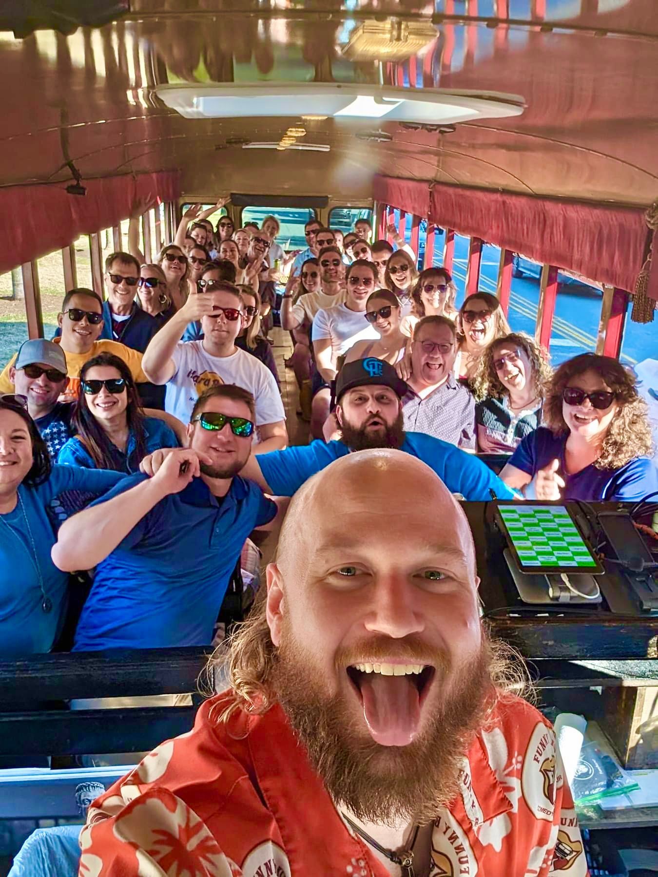 A man with a beard and orange patterned shirt smiles for a selfie on a crowded bus with many passengers behind him.