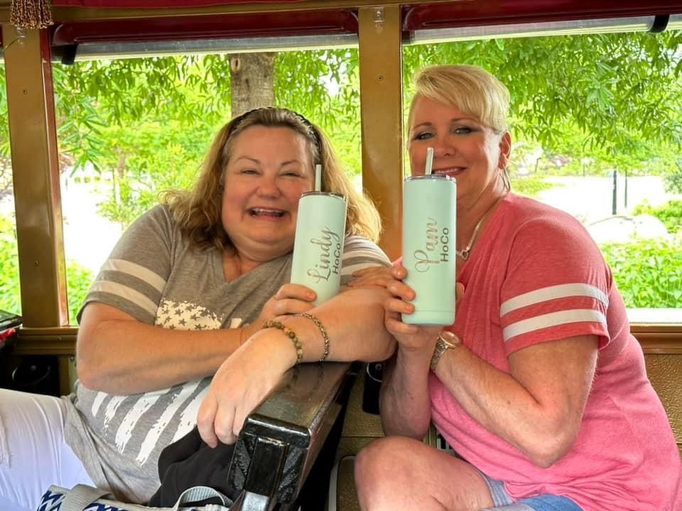 Two people smile and hold up matching light blue tumblers while sitting in an outdoor, covered area with greenery nearby.