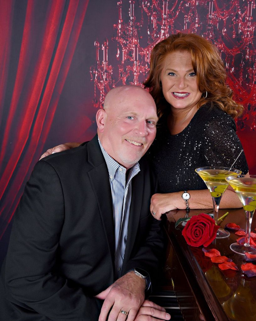 Woman in black dress and man in blue shirt pose together, smiling, against a dark gray background.