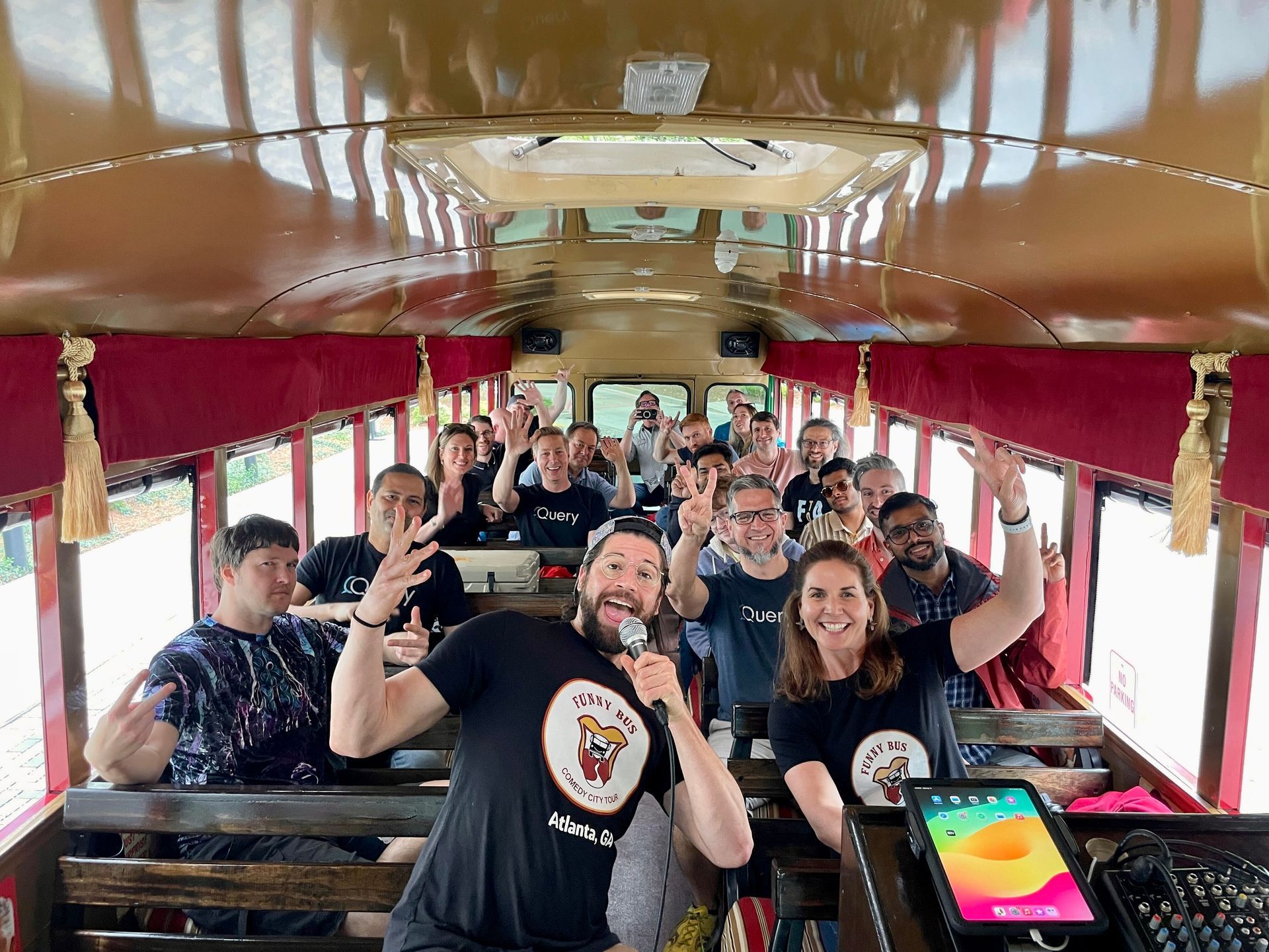 A group of people smiles and gestures on a wooden-interior bus, with a man in the foreground holding a microphone.