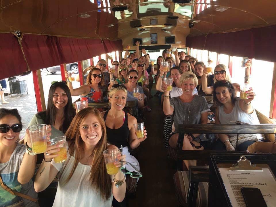 A group of people on a trolley ride indoors, smiling and holding up plastic cups for a toast.