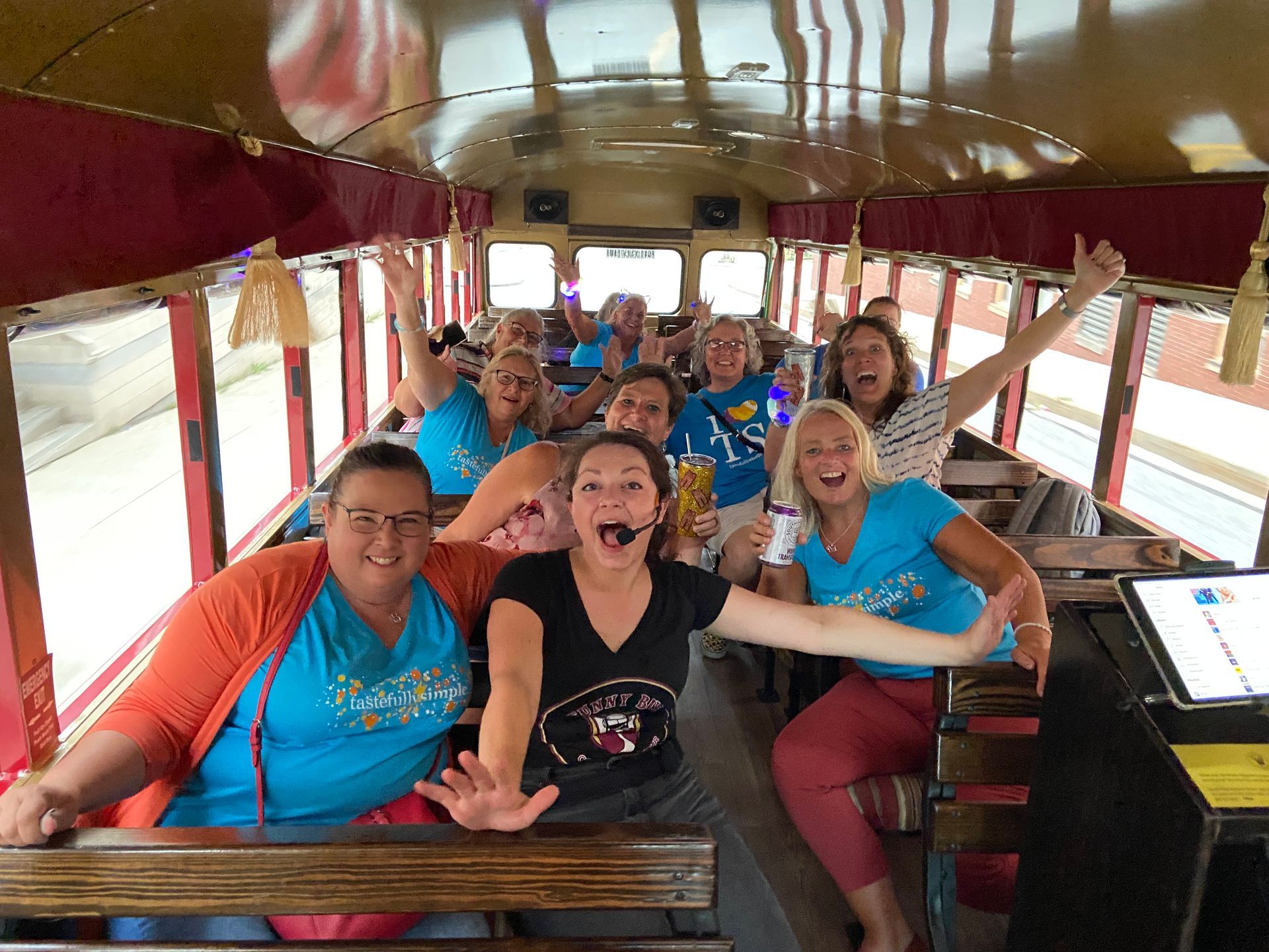 Group of people on a decorated trolley, smiling and wearing Christmas lights, some holding drinks.