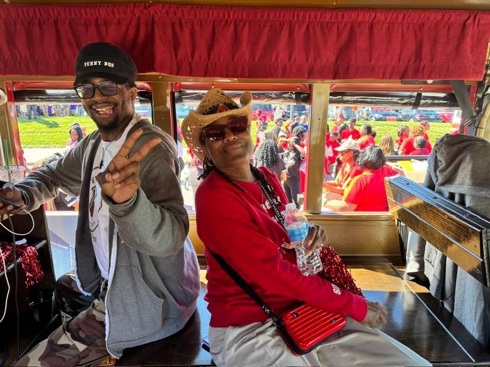 Two people smile and pose inside a trolley on a sunny day; one person makes a peace sign while wearing a cap and glasses.