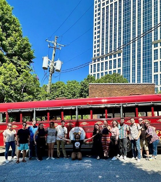 Group posing in front of a red trolley under a blue sky with tall buildings and trees nearby